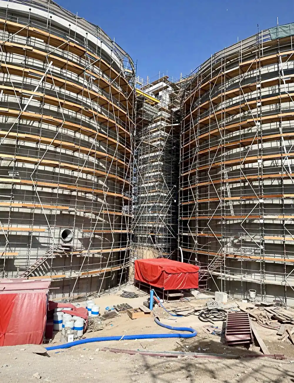 Low-angle view looking up between two large digester tanks, highlighting the extensive scaffolding and the challenging elevated work environment.